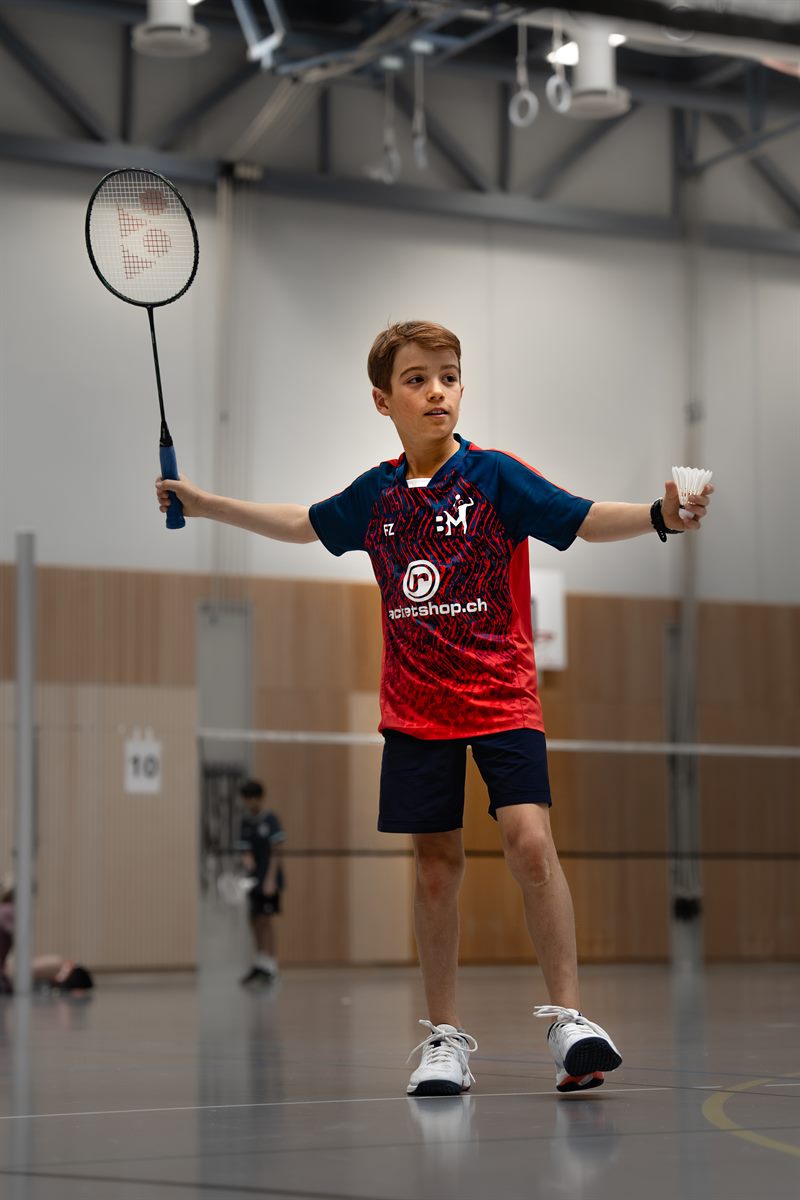 Junger Badmintonspieler im roten Trikot hält Schläger und Federball und bereitet den Aufschlag vor