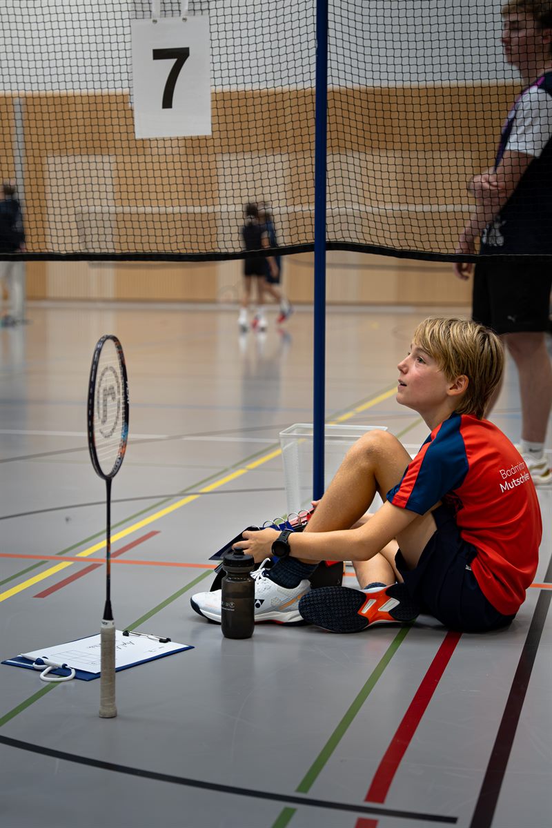 Junger Spieler sitzt in der Pause am Spielfeldrand neben Schläger und Trinkflasche, dahinter Feld 7