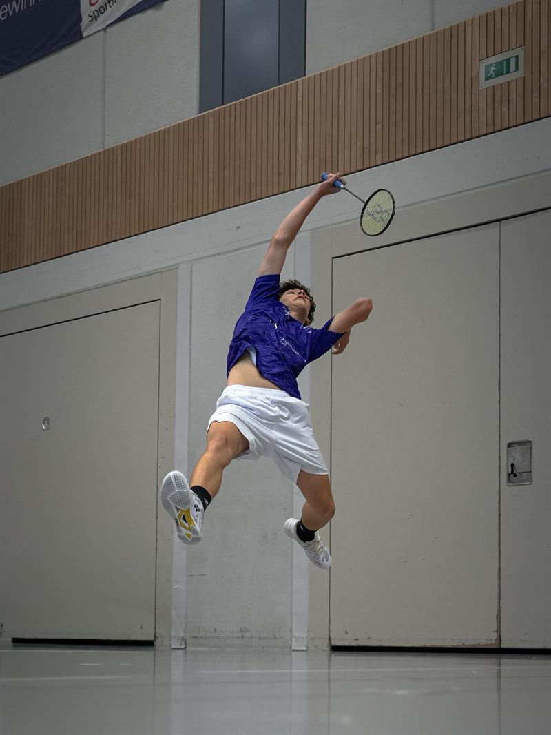 Badmintonspieler im violetten Trikot beim Sprungsmash, der Schläger weit über dem Kopf, vor heller Hallenwand