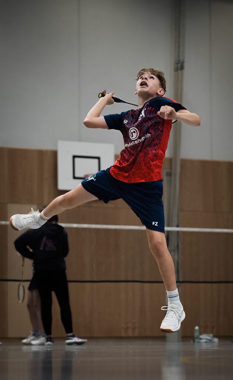 Junger Badmintonspieler im roten Trikot springt zum Schlag, den Schläger hinter dem Kopf, in einer Sporthalle