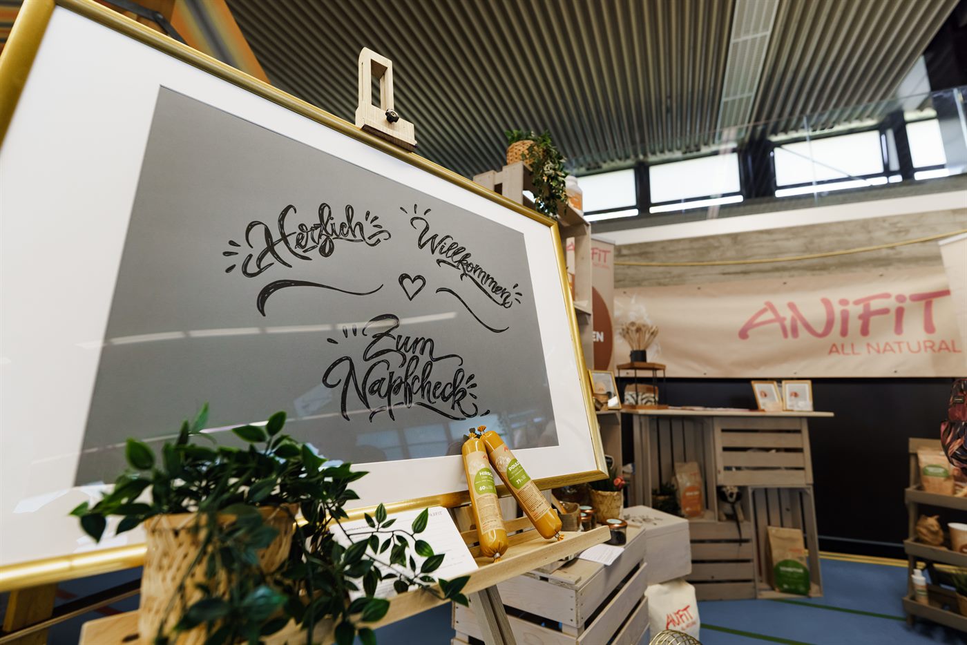 Tafel mit handgeschriebenem Willkommensgruss an einem Messestand, daneben eine Pflanze