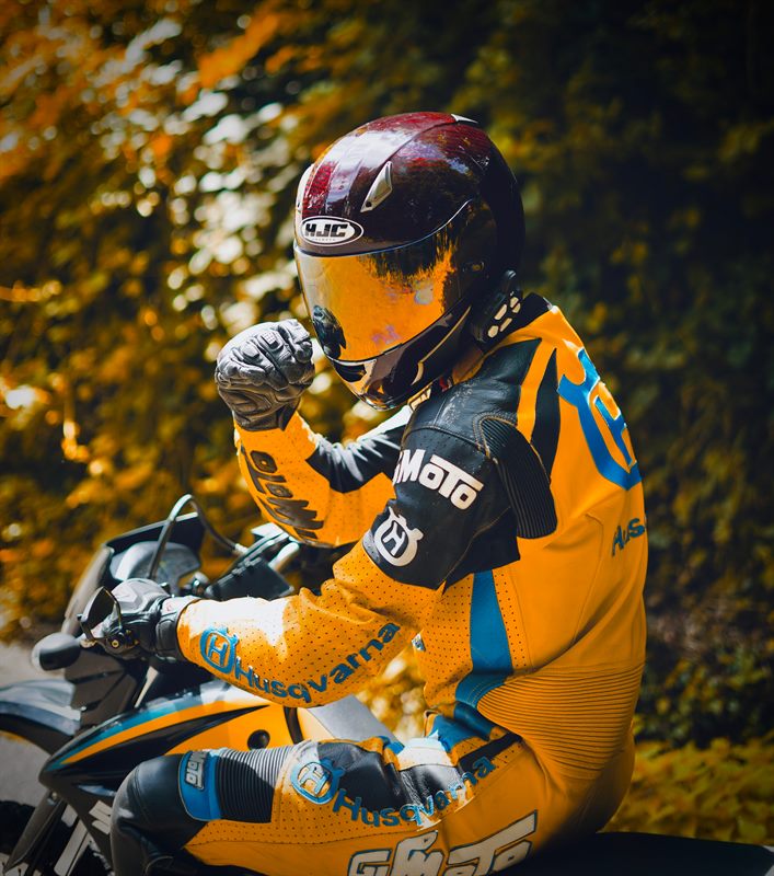 Motorradfahrer in gelb-schwarzer Lederkombi und Helm stützt sich auf sein Motorrad, warmes Herbstlaub im Hintergrund