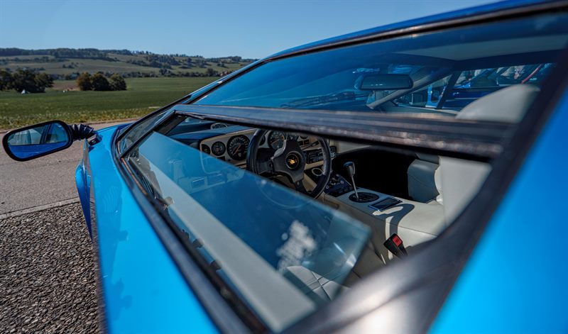 Blick durch das Seitenfenster eines blauen Lamborghini ins Cockpit mit Stier-Logo auf dem Lenkrad