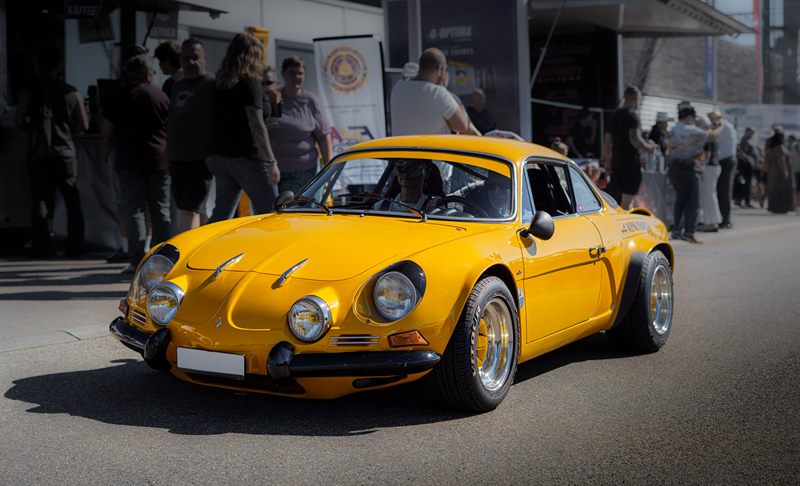 Gelber klassischer Sportwagen von vorne an einem Autotreffen, dahinter Besucher