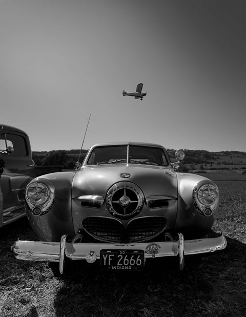 Studebaker der 1950er-Jahre von vorne auf einer Wiese, darüber ein Kleinflugzeug, schwarz-weiss